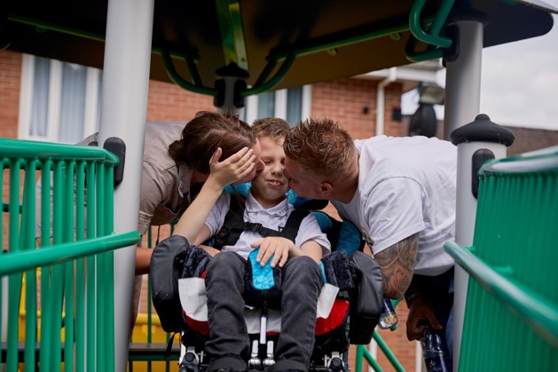Lucas is sitting in his wheelchair. He is smiling. His mum Becci and dad Andy are kissing his checks on each side.