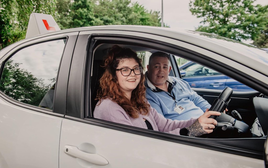 Imogen is sitting in the driving seat of her instructors car, it has a learner plate on top of the vehicle. She has light brown hair, black glasses, lilac jumper, black tattoos on her right hand and she is smiling. Her instructor is in the passenger seat. He has short dark hear and a light blue polo top on. 