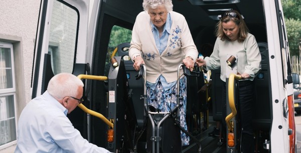 Lisa is standing in the back of a minibus with the doors open, she is supporting an older women who is stepping onto a lift at the back of the vehicle with her mobility aid. A gentleman is assisting the older women by controlling the lift.