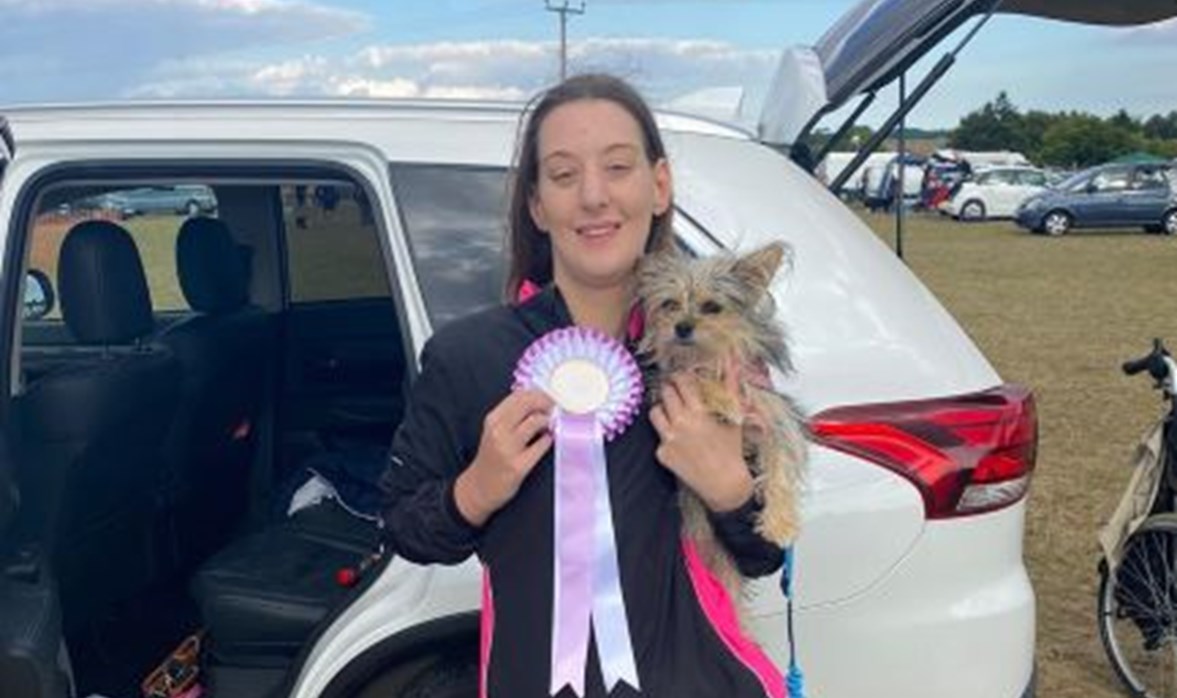 A young women is smiling holding a small dog in one hand and a rosette in another.  