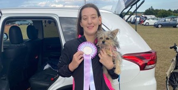 A young women is smiling holding a small dog in one hand and a rosette in another.  