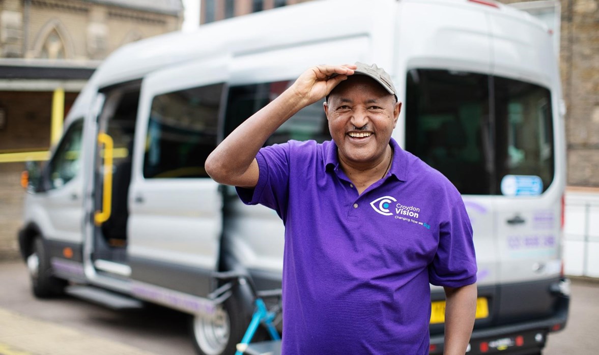 A gentleman is standing in from of a community transport vehicle smiling. 