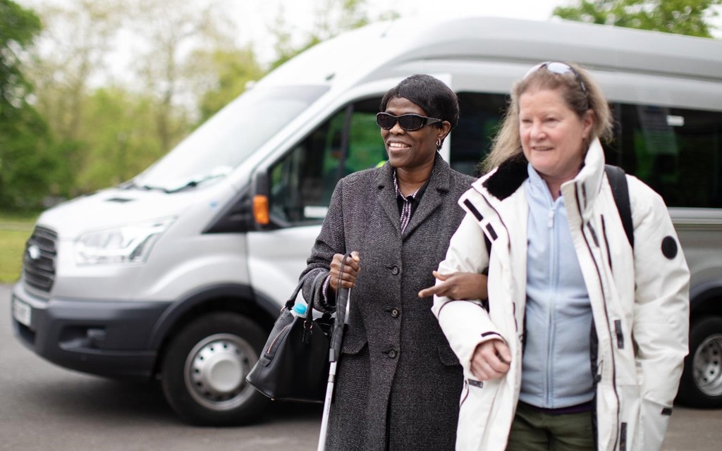 A women has linked arms with another woman as they walk away from a silver minibus which sits in the background. The first women is smiling, she is wearing a wool coat, sun glasses, black handbag and is using a walking aid. The second women has a white coat on and a light blue fleece underneath with sunglasses on her head. 