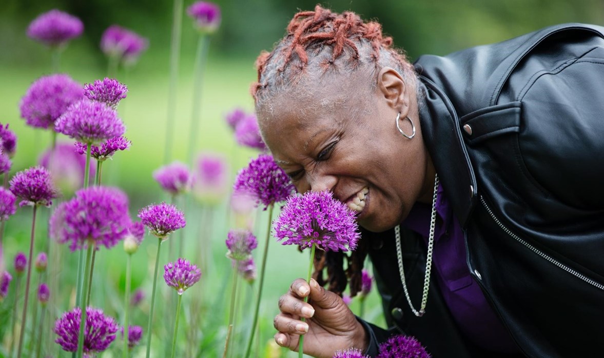 A lady in a black leather jacket and purple polo is smelling a bright purple flower. 