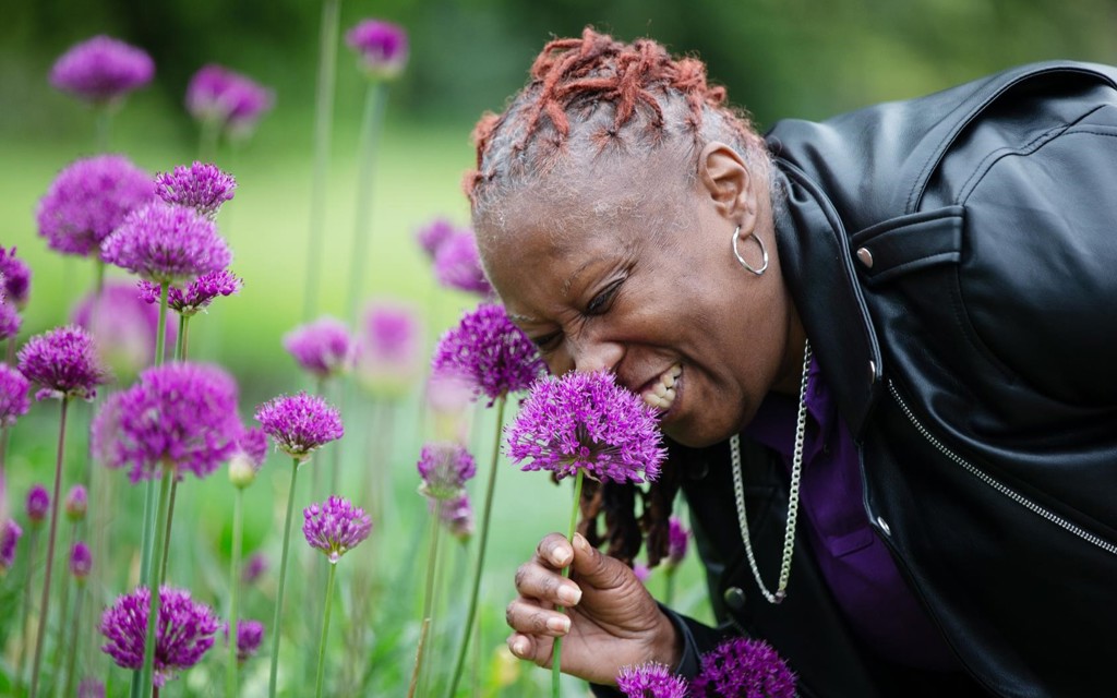 A lady in a black leather jacket and purple polo is smelling a bright purple flower. 