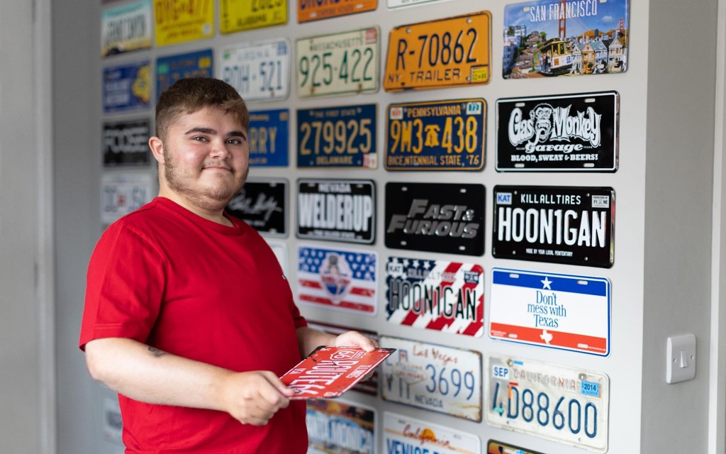 Harry is a young man standing in front of a wall with car number plates smiling. 