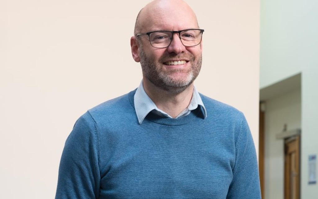 Nigel Fletcher CEO is smiling in a blue jumper with black rimmed glasses. 