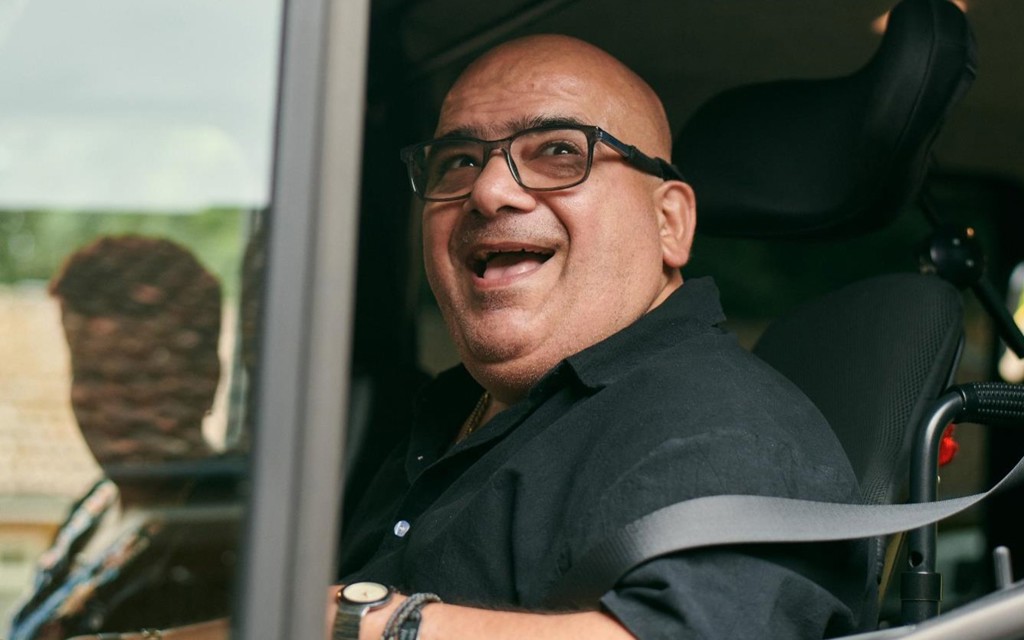 Pete smiling while sitting in his vehicle, the window is down and Angela can be seen in the reflection of the car window. 