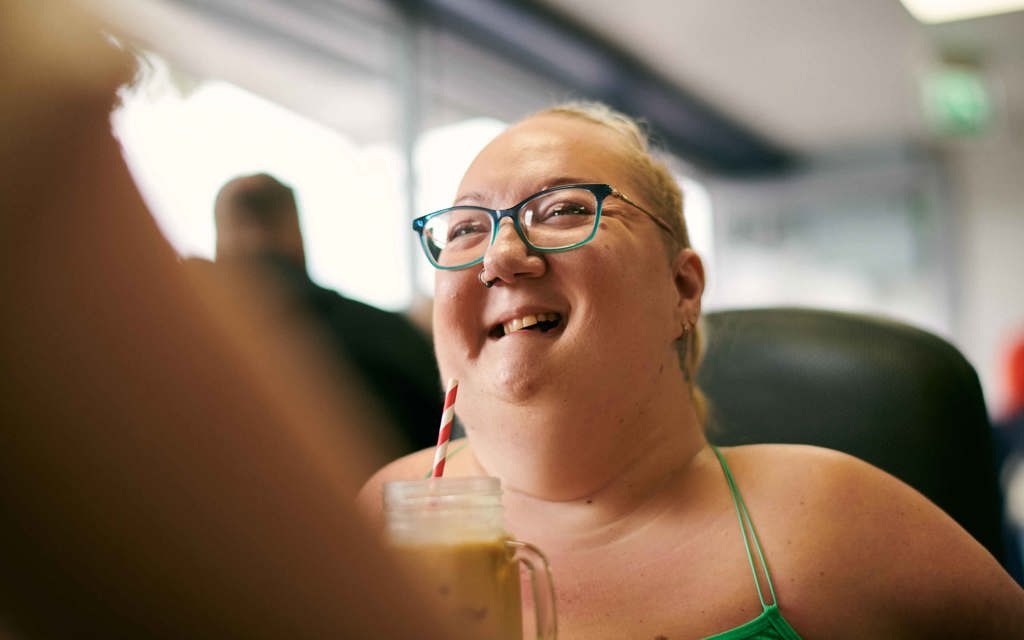 Tina is in a café. She is smiling and talking to her friend.