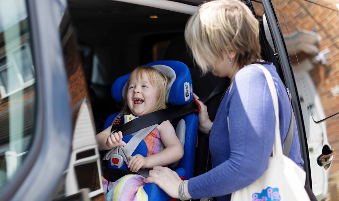 Poppy's car door is slid open. Poppy has a big smile on her face. Her mum is carefully strapping her into car seat before they take a trip out for the day.