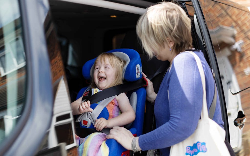 Poppy's car door is slid open. Poppy has a big smile on her face. Her mum is carefully strapping her into car seat before they take a trip out for the day.