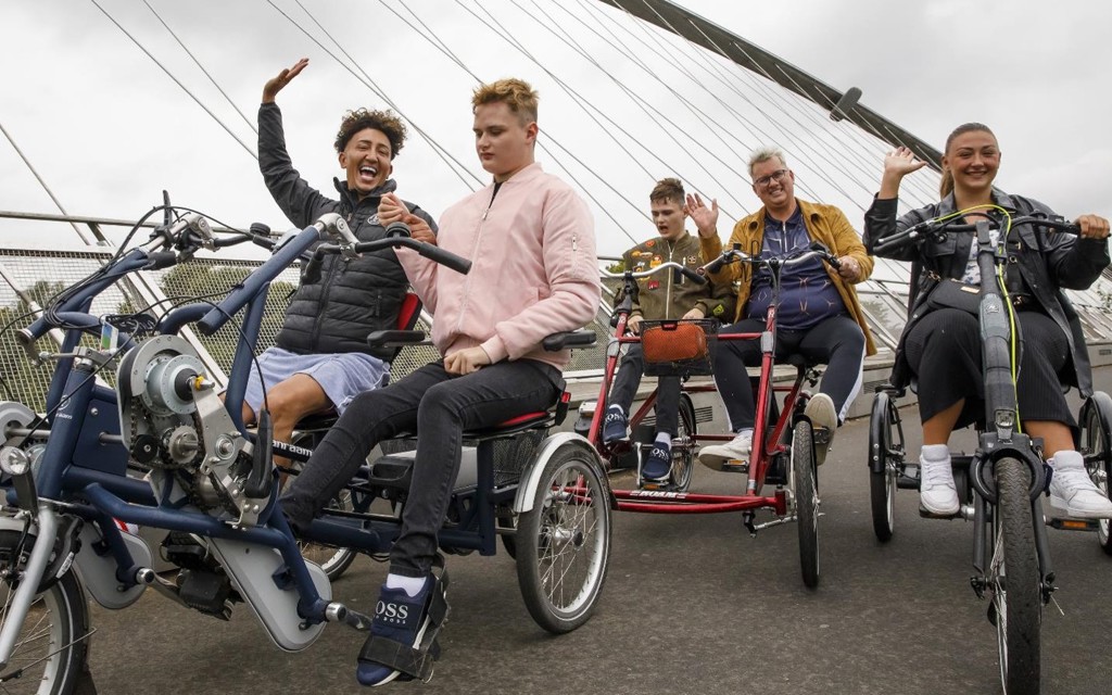 Three inclusive cycles are pictured on a bridge. The first seats two young men one has brown hair, black coat, and blue shorts, the other has blond hair, pink jacket, black jeans, and black trainers. Another cycle has a young man with brown hair in a green jacket with badges, black trousers and blue trainers and a man with white hair, yellow jacket, blue jumper, black trousers, and white trainers. The final cycle is a one-person cycle, a young woman is sat with brown long hair, black jacket, black trousers.