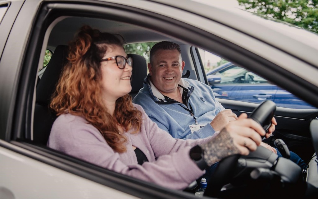 Imogen is sitting in the driving seat of her instructors car, it has a learner plate on top of the vehicle. She has light brown hair, black glasses, lilac jumper, black tattoos on her right hand and she is smiling. Her instructor is in the passenger seat. He has short dark hear and a light blue polo top on. 