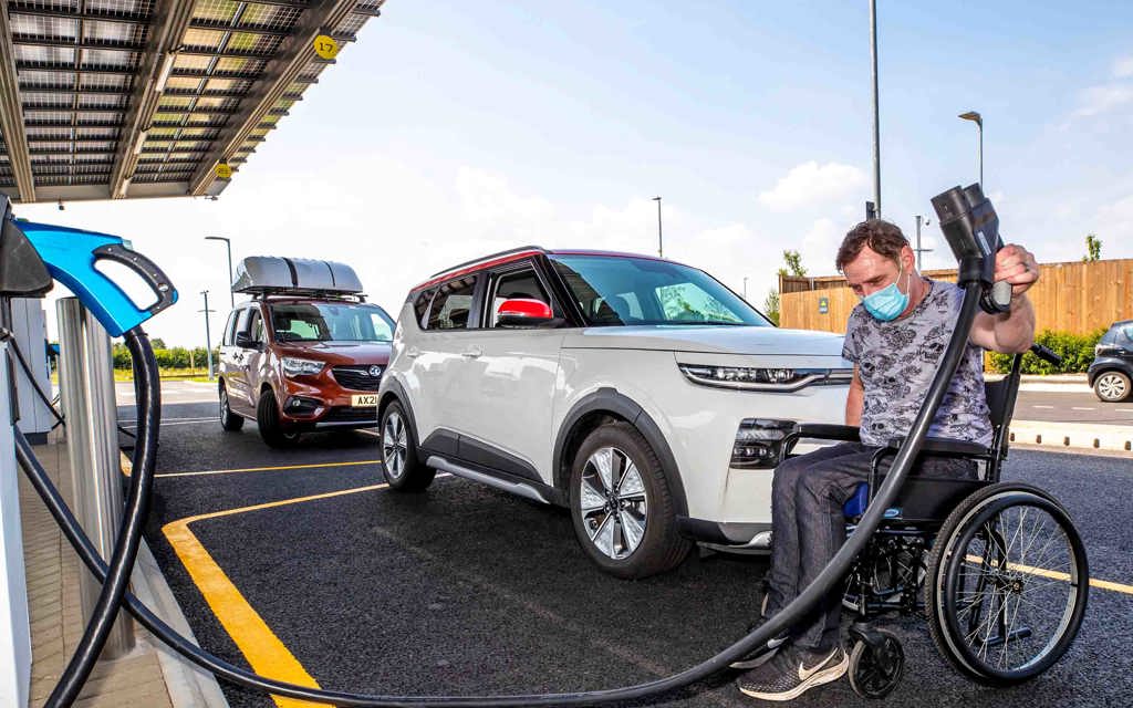 A man in a wheelchair is holding a charging plug for an electric car. The wheelchair is in front of a parked electric vehicle.