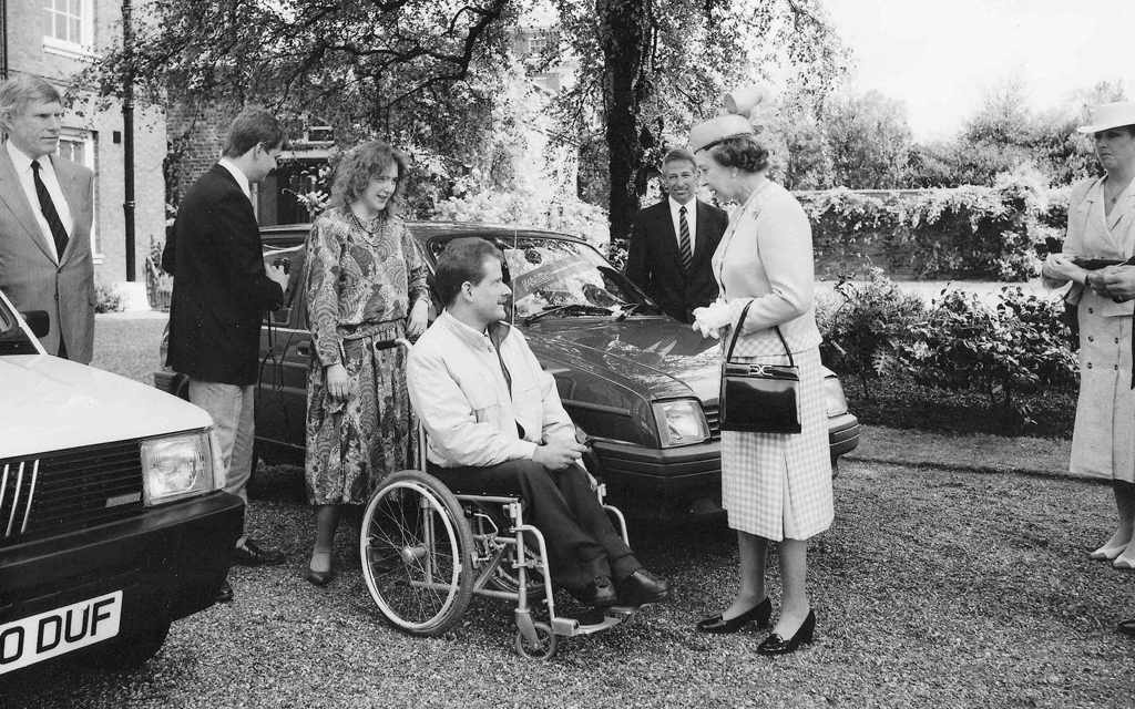 Her Majesty The Queen standing in front of a car and talking to a gentleman who is sitting in a wheelchair. They are both smiling.