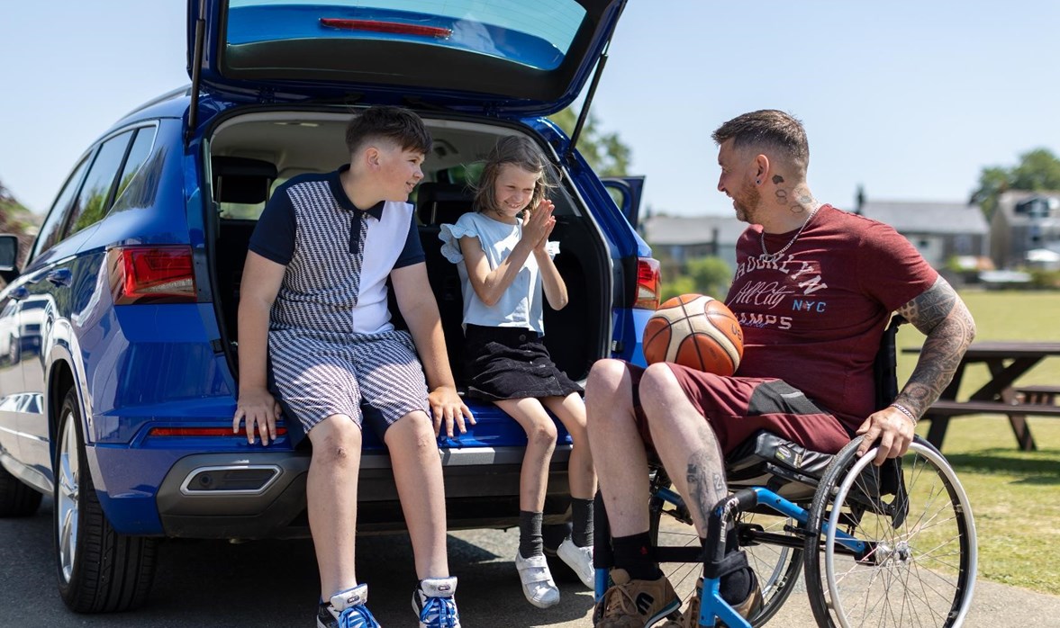 Aaron is sitting in his wheelchair next to his car. The car boot is open and his children are sitting in it. They are all talking to one another.