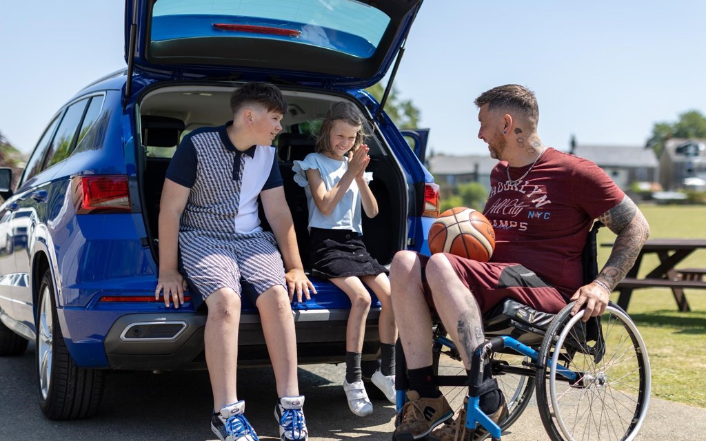 Aaron is sitting in his wheelchair next to his car. The car boot is open and his children are sitting in it. They are all talking to one another.