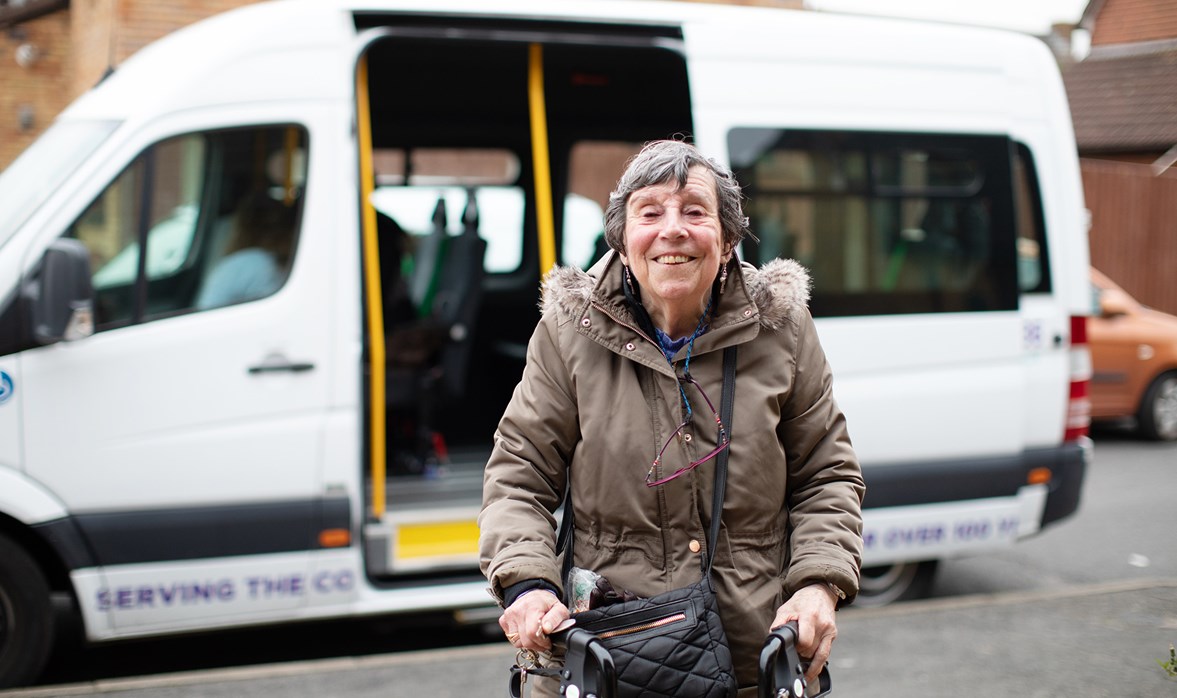 A smiling woman with a walking frame stands outside in front of a minibus