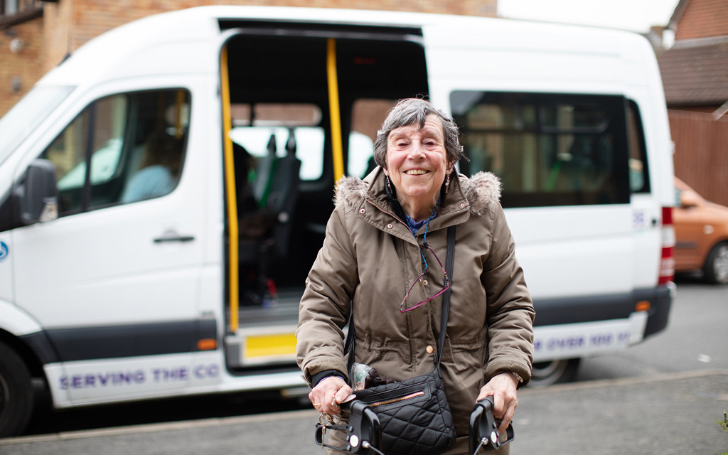 A smiling woman with a walking frame stands outside in front of a minibus