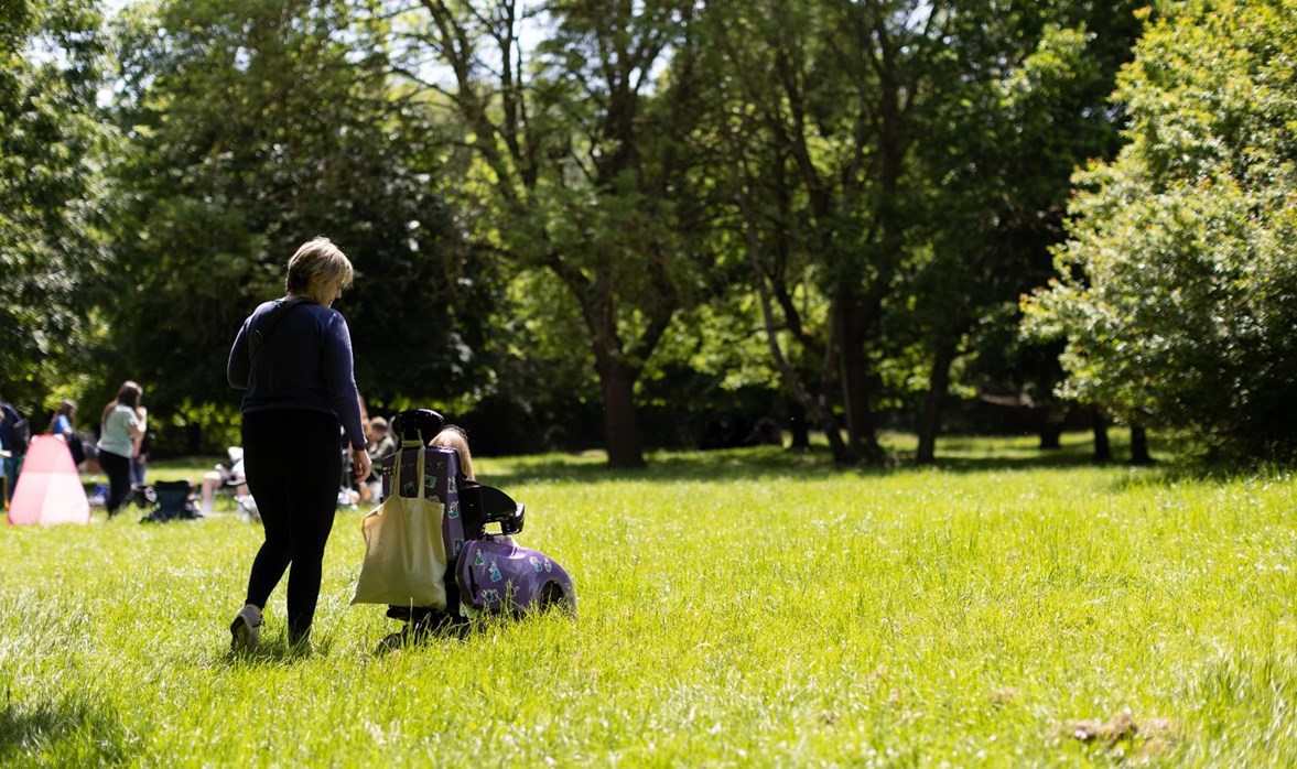 A young girl is in a light purple coloured wheelchair with a lady standing next to her walking through a lightly lit grassy field. 