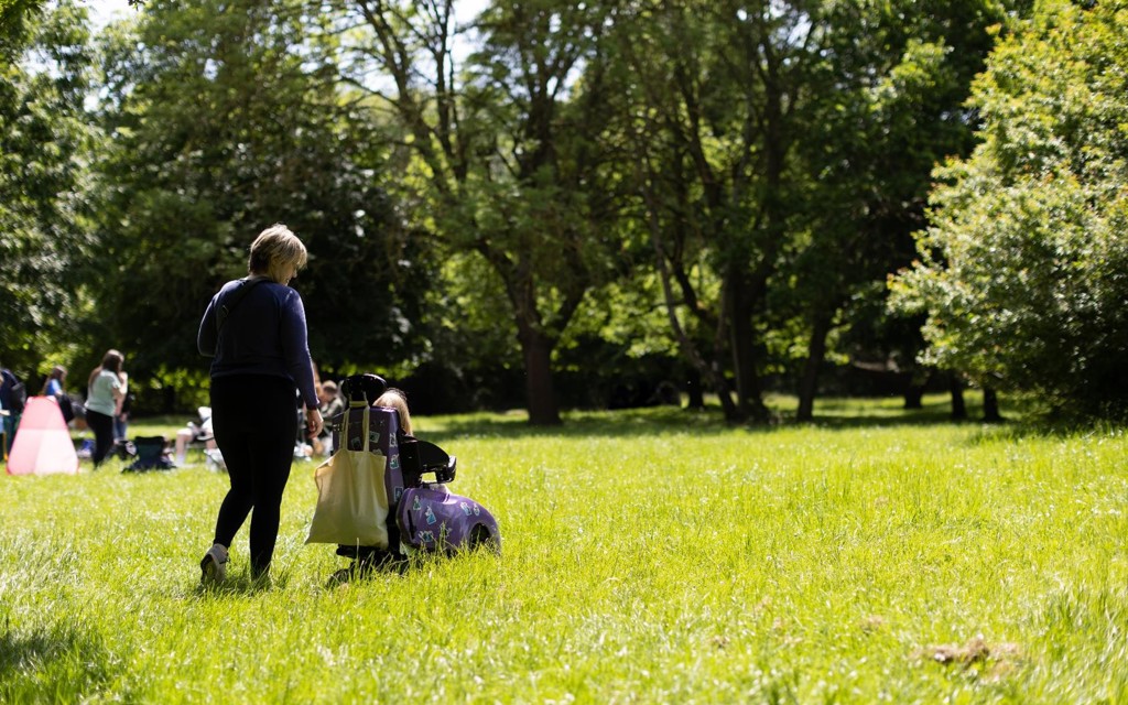 A young girl is in a light purple coloured wheelchair with a lady standing next to her walking through a lightly lit grassy field. 