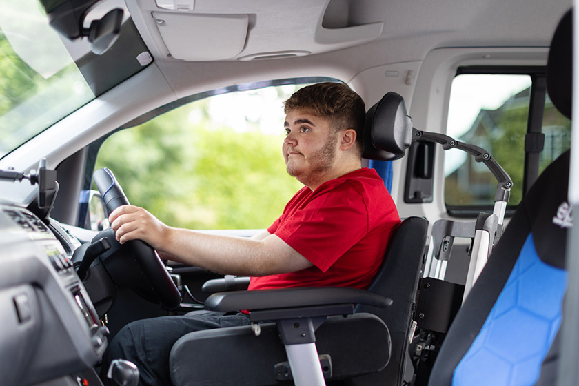 Harry is looking in the back mirror of his vehicle smiling. He is in his powered wheelchair at the steering wheel of his vehicle.