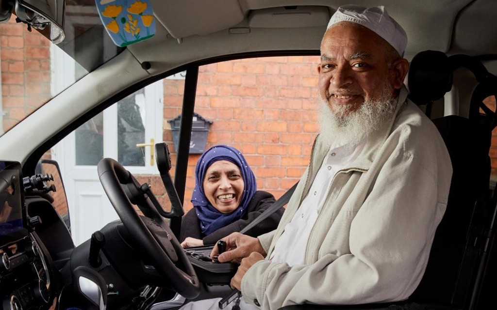 Moosa is sitting in his vehicle with hand controls smiling. His wife is behind the car, standing next to Moosa, smiling. 