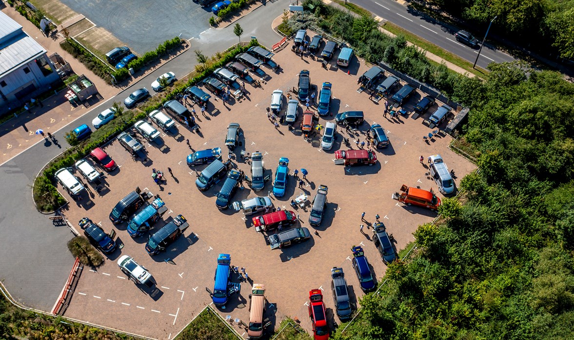 An image of vehicle parked in a carpark and people walking around looking at the vehicles from above. 