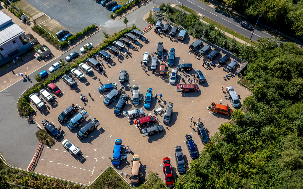 An image of vehicle parked in a carpark and people walking around looking at the vehicles from above. 