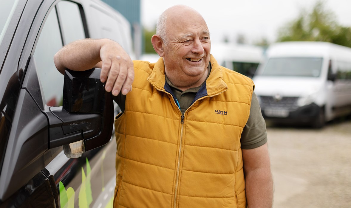 A smiling man wearing a yellow vest stands next to a wheelchair accessible vehicle with his arm placed on the mirror