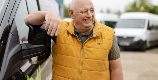 A smiling man wearing a yellow vest stands next to a wheelchair accessible vehicle with his arm placed on the mirror