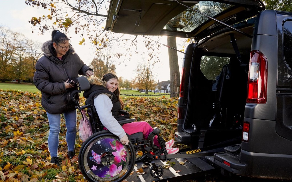 Becky is in her wheelchair on the ramp of her black wheelchair accessible vehicle. She is smiling to camera. She has long brown hair, black coat, grey hoodie, pink trousers and pink trainers. He mum Sarah is operating the ramp, she is has black long hair in a pony tail with a black coat, blue jeans and black trainers, she is smiling at Becky. Behind them is a park with orange and yellow leaves on the trees.