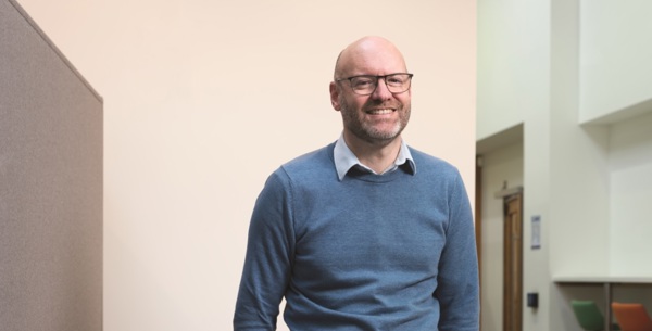 Nigel Fletcher CEO is smiling in a blue jumper with black rimmed glasses.