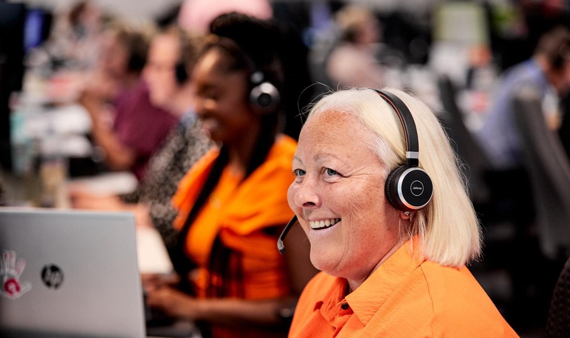 A lady in a bright orange is answering the telephone using a headset and smiling.  