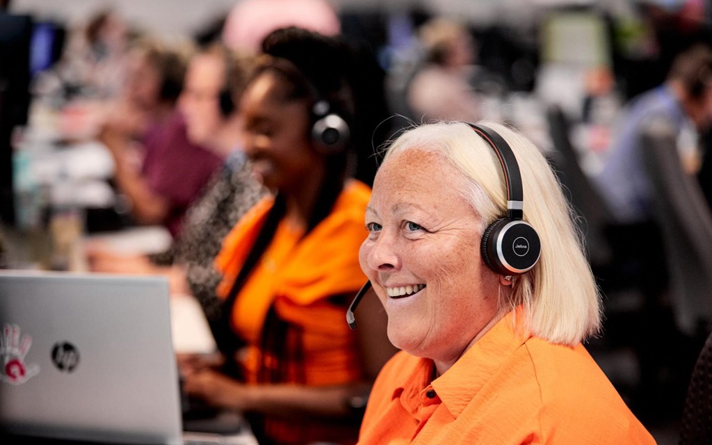 A lady in a bright orange is answering the telephone using a headset and smiling.  