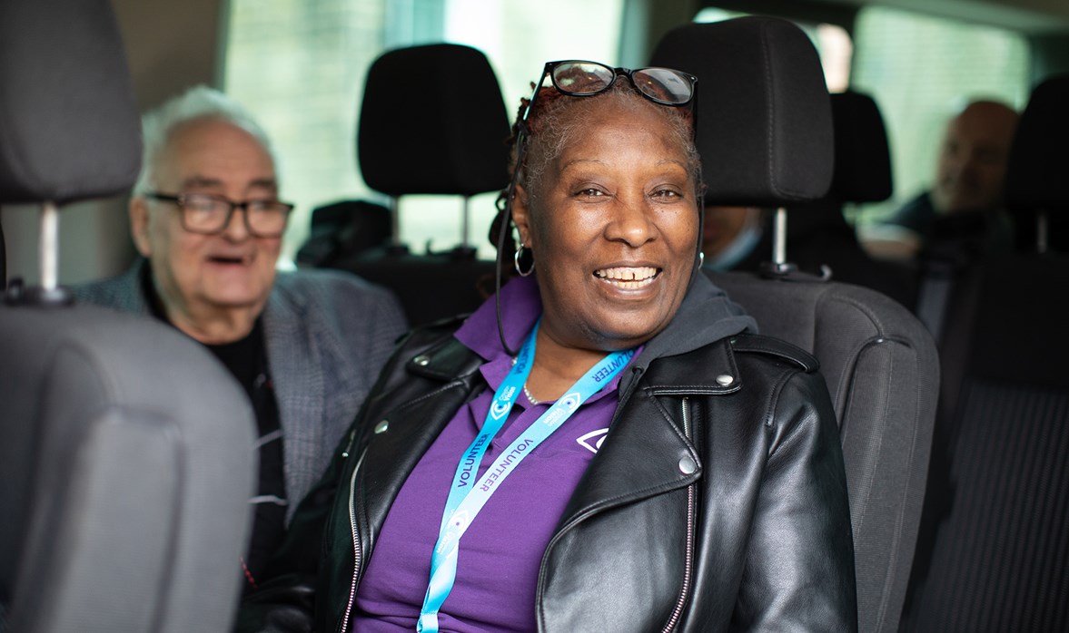 A smiling woman wearing a purple polo shirt sits in a minibus next to a man