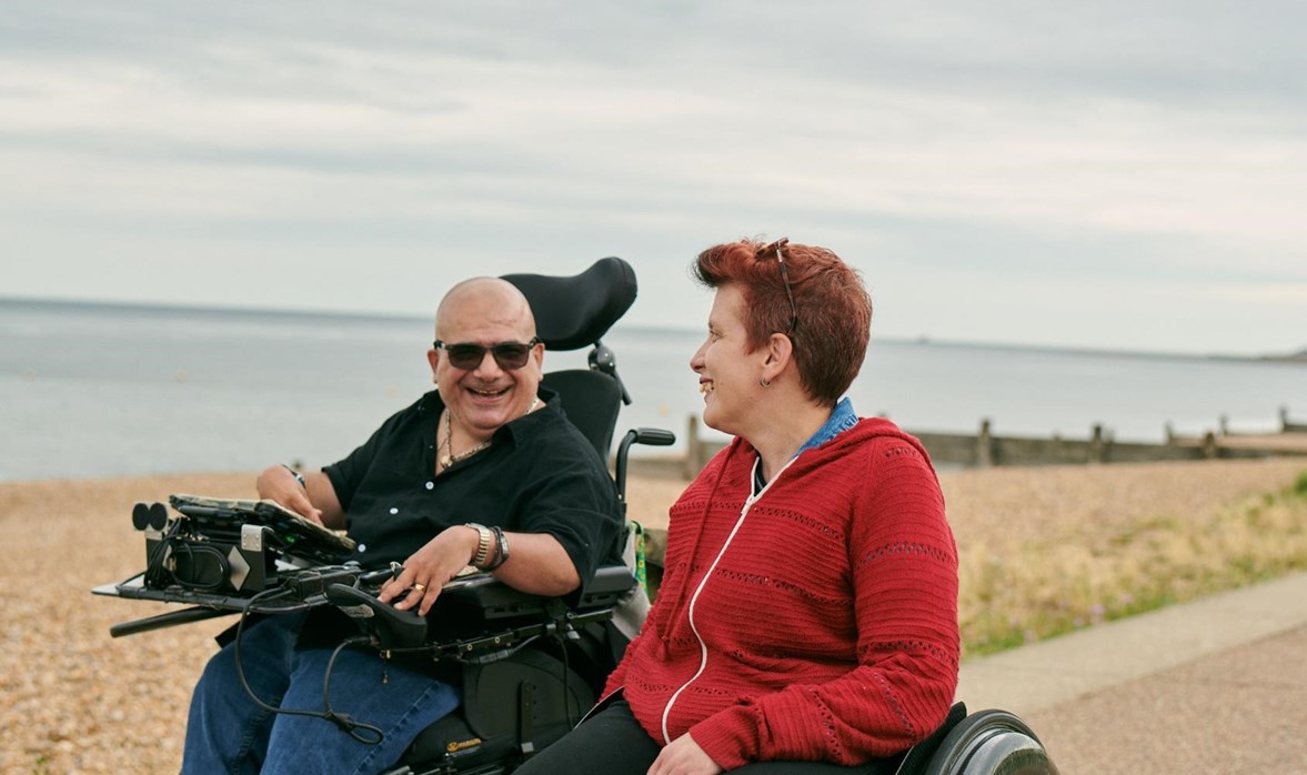 Angela and Pete are wheeling along the sea side, smiling and talking. 