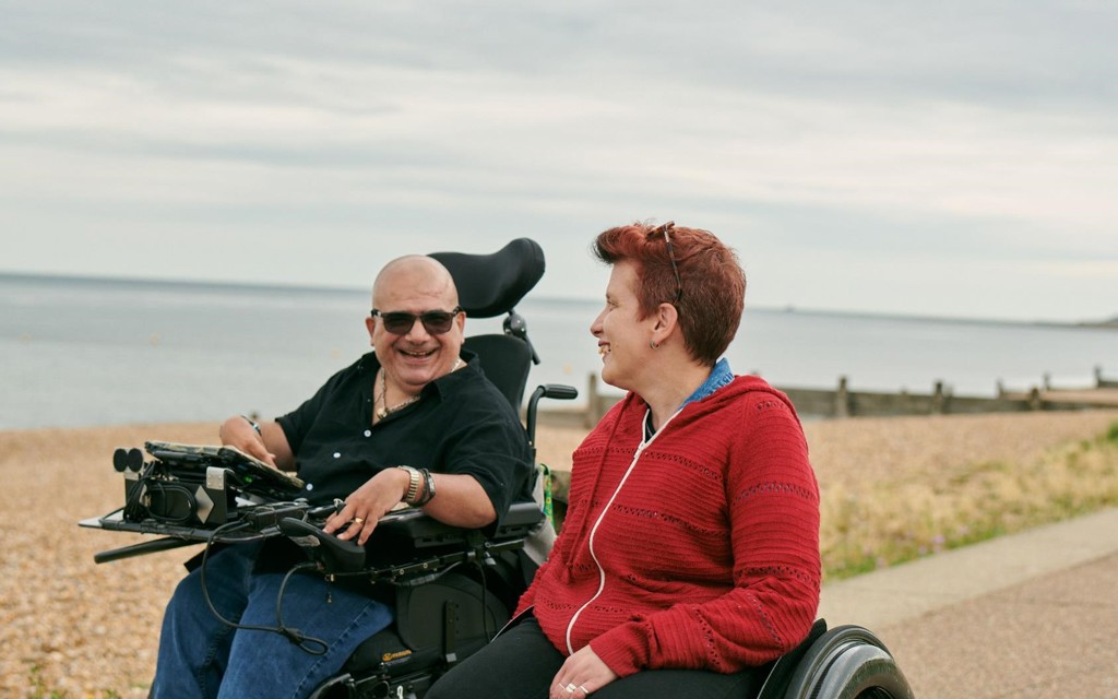 Angela and Pete are wheeling along the sea side, smiling and talking. 