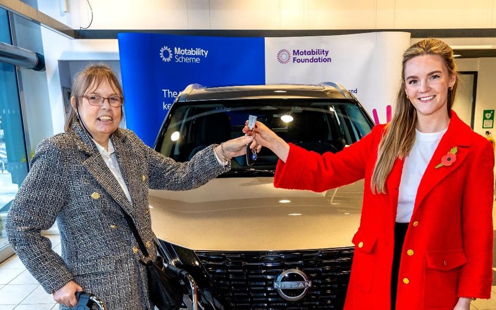 The picture has been taken in Bristol Street Motors Nissan, Halifax. Kate Dearden MP is presenting car keys to Allison. There is a silver car parked behind them.