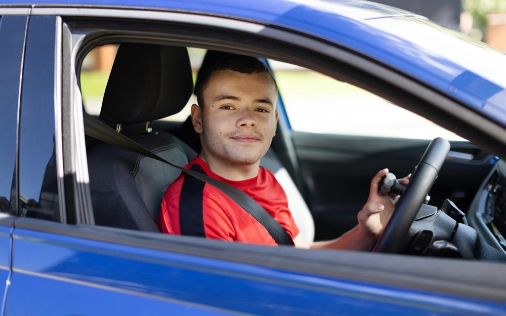 A young man is sitting and smiling in the front driving seat of his blue vehicle. He is wearing a red top with brown short hair.