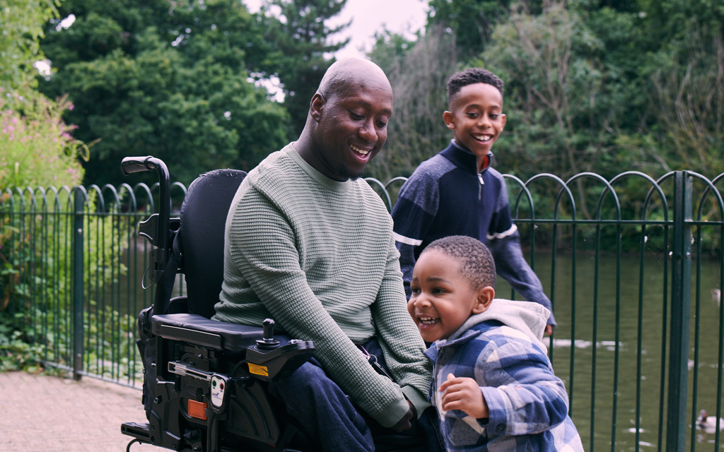 A man in a wheelchair plays with his two sons beside a lake in a park