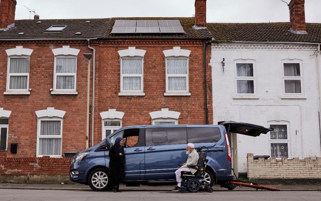 Moosa is siting in his electric wheelchair with his blue wheelchair accessible vehicle behind, the wheelchair ramp is down. His wife is standing opposite him, they are both smiling. 