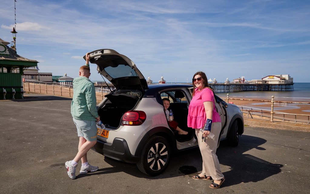 Jess is outside at the beach, standing next to her car smiling. Her family are with her. 