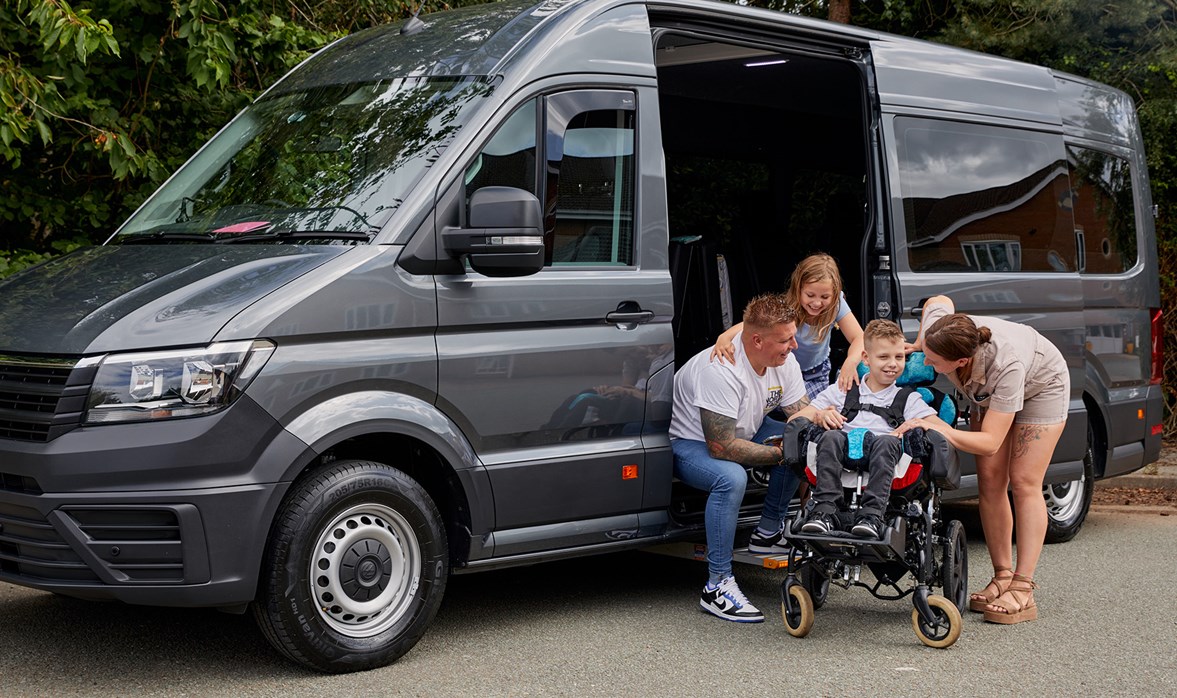 A family sit next to their grey wheelchair accessible vehicle