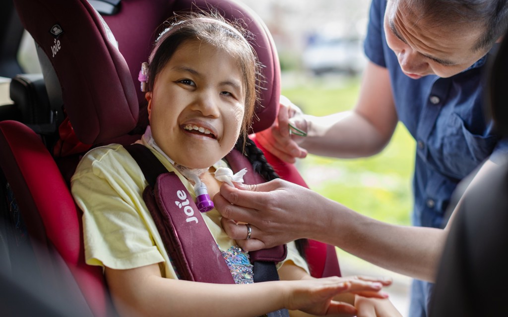 Eva is smiling at the camera in a yellow top, pink headband, she is sitting in her bright pink car seat. Eva's dad is helping her into the car seat smiling. He is in a navy blue button up short sleeved shirt.