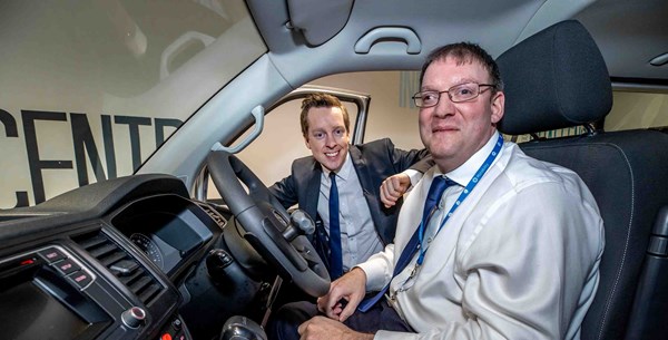 The photo shows the inside of an adapted vehicle. Sat in the front seat of the vehicle is a Motability employee smiling with his hand on knees, wearing a smart suit. Tom Pursglove MP is smiling and looking in the vehicle from outside next to the employee with his arm rested on the open right front door.