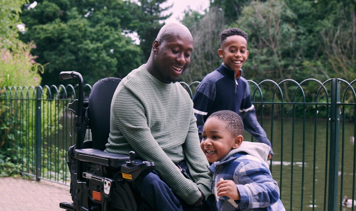 A gentleman is smiling in a powered wheelchair with his two sons next to a pond at the park. 