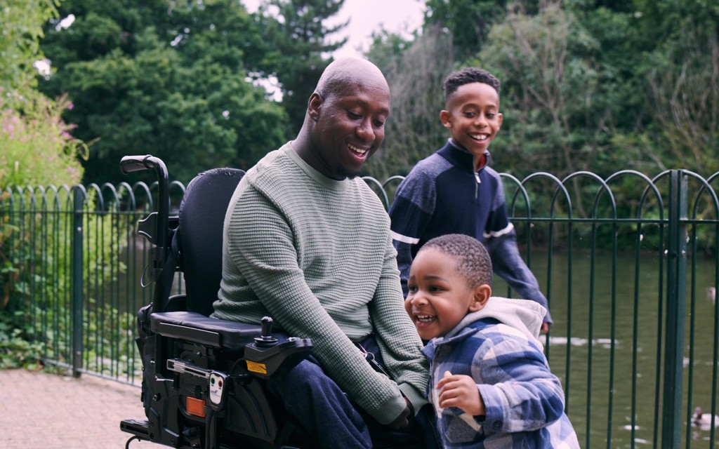 A gentleman is smiling in a powered wheelchair with his two sons next to a pond at the park. 