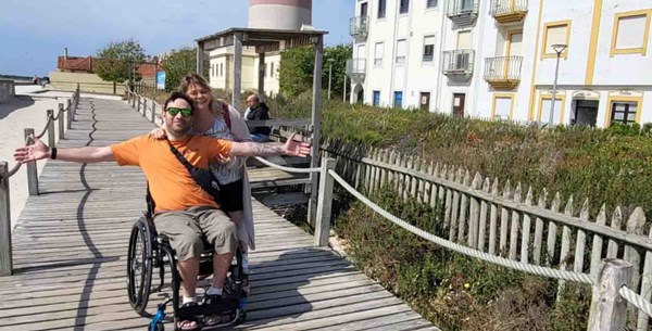 Pedro is sitting in his wheelchair smiling with his arms open. A lady is laughing with an arm around his shoulder. They are at the beach, enjoying the sun.