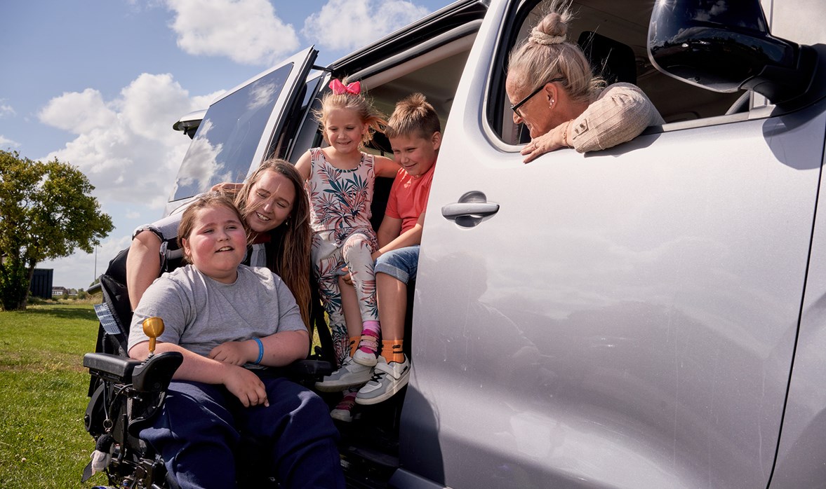 A boy sits in a powered wheelchair alongside his family next to their wheelchair accessible vehicle.
